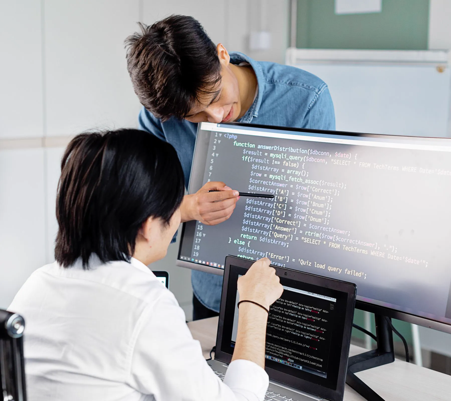 A senior engineer walking a colleague through code on a large monitor in an office